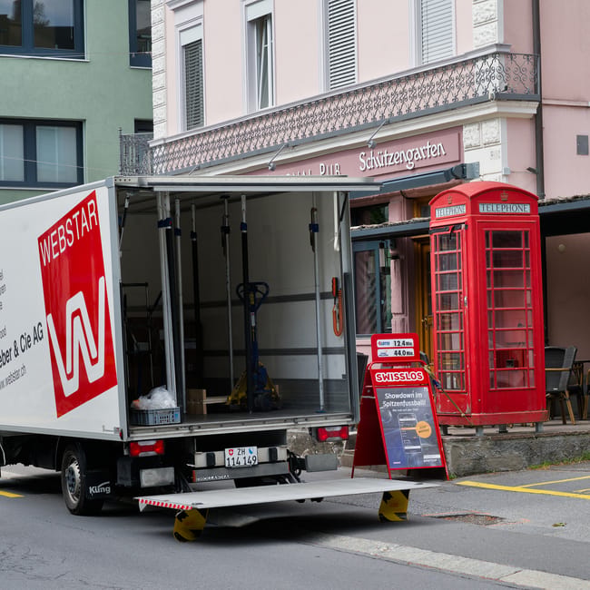 A dark red flatbed van parked on a city street with a modern office building in the background. The vehicle's bed is loaded with various household items, including several large cardboard boxes wrappe