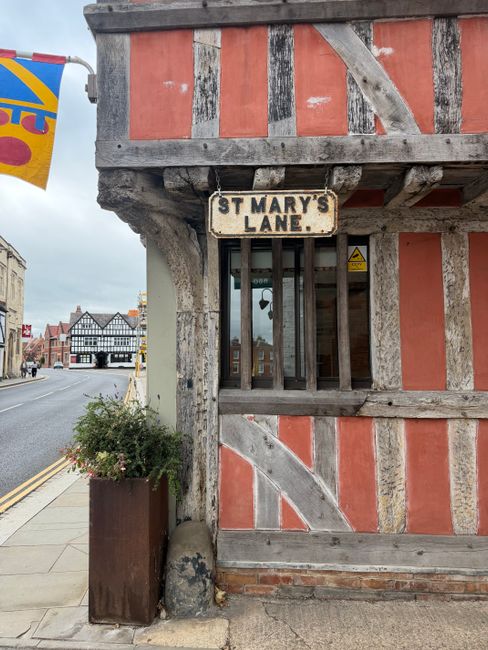 A close-up view of a traditional timber-framed building on St Mary's Lane, Upminster, showing weathered wooden beams and orange-colored infill panels. A small, barred window is located beneath a woode