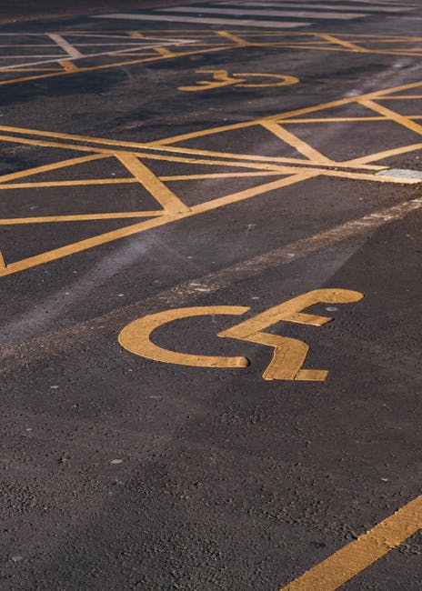 A close-up view of a designated accessible parking space in an outdoor parking lot, with the yellow wheelchair symbol painted on the dark asphalt surface. The parking area features multiple parking bays separated by yellow lines, some of which are partially visible in the background. The surface shows signs of wear and slight smoothing where vehicles have moved, with areas of minor discoloration. The image is taken during daylight, with natural light illuminating the scene. This setting relates to the process of home relocation and the need for accessible parking permits during moving and furniture transport, which services like Man with Van Upminster may assist with in Upminster and Havering Council areas.