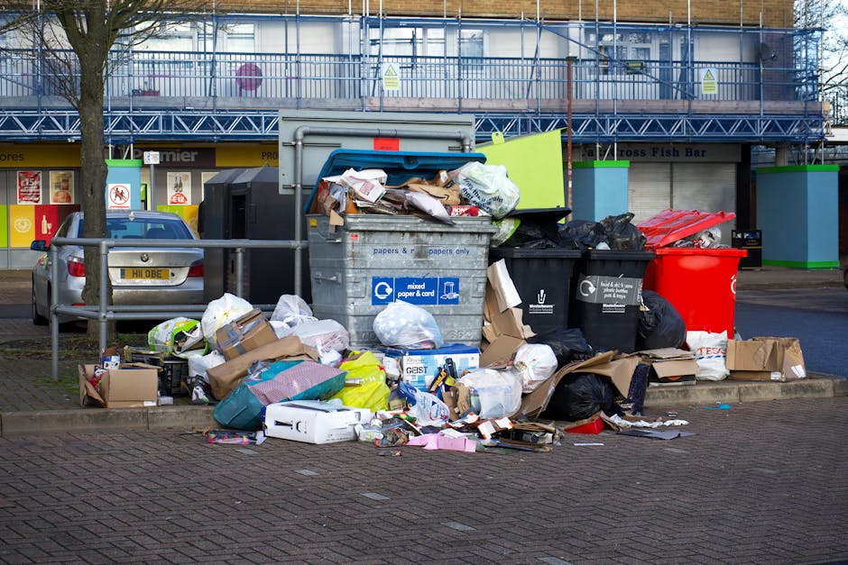 A large pile of mixed waste and household rubbish with partially open cardboard boxes, plastic bags, and paper and cardboard recyclables spilling onto a paved sidewalk in front of a recycling collection point. The collection point includes several large bins and containers, such as a gray mixed paper and cardboard bin, a black general waste bin, and a red recycling bin. Behind the bins, a metal fence separates the area from a commercial building with signage and storefronts, including a fish bar. A silver hatchback car is parked nearby, and there are tall, leafless trees and a blue scaffold structure in the background. The scene is illuminated by daylight, and this image relates to household waste disposal, often associated with home relocation or moving processes, as demonstrated by the presence of various packing materials and transportation equipment occasionally used by companies like Man with Van Upminster during removals or furniture transport.