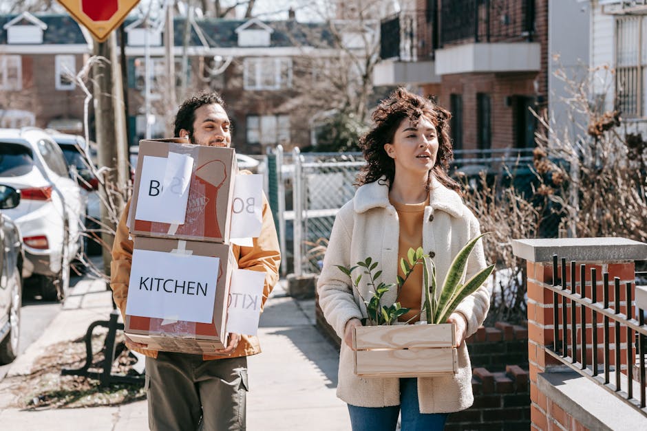 Two individuals are depicted outdoors during daytime, engaged in a house removal process. The man is carrying a medium-sized cardboard box labeled 'KITCHEN' with additional packing papers attached and is wearing a brown jacket and beige trousers. He is smiling and walking on a paved sidewalk near parked cars and residential buildings. The woman, walking ahead, is dressed in a white fleece coat over a brown top, and is holding a wooden crate filled with potted plants, including a snake plant and other greenery. They are approaching a brick and metal fence, suggestive of an entryway to a property where furniture and belongings are likely being transported. Visible moving equipment such as trolleys and blankets are not present in the image, but the scene implies an organized home relocation effort. The background includes leafless bushes and multi-storey residential buildings, set in a suburban neighbourhood. This image illustrates the physical process of packing, carrying, and transporting household items, aligned with services offered by Man with Van Upminster during home moves, furniture transport, and packing and moving tasks related to relocating to Corbets Tey Estate, Upminster.