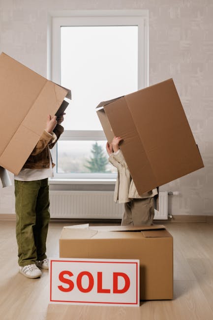 Two individuals are depicted outdoors during daytime, engaged in a house removal process. The man is carrying a medium-sized cardboard box labeled 'KITCHEN' with additional packing papers attached and is wearing a brown jacket and beige trousers. He is smiling and walking on a paved sidewalk near parked cars and residential buildings. The woman, walking ahead, is dressed in a white fleece coat over a brown top, and is holding a wooden crate filled with potted plants, including a snake plant and other greenery. They are approaching a brick and metal fence, suggestive of an entryway to a property where furniture and belongings are likely being transported. Visible moving equipment such as trolleys and blankets are not present in the image, but the scene implies an organized home relocation effort. The background includes leafless bushes and multi-storey residential buildings, set in a suburban neighbourhood. This image illustrates the physical process of packing, carrying, and transporting household items, aligned with services offered by Man with Van Upminster during home moves, furniture transport, and packing and moving tasks related to relocating to Corbets Tey Estate, Upminster.