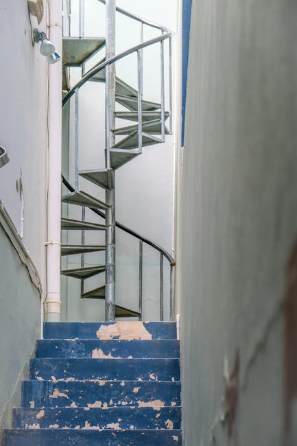 Interior view of a narrow staircase with worn blue-painted steps featuring peeling paint, leading upwards within a residential property. The staircase is enclosed by a pale green wall on the right and a white wall with visible pipes on the left. At the top of the stairs, a small section of a spiral metal staircase with steps made of metal and a handrail is visible, indicating a multi-level home. The environment appears aged, with signs of wear and tear on surfaces. In the context of house removals, this scene illustrates the challenges of navigating narrow, Victorian-era staircases during furniture transport or packing and moving processes, which companies like Man with Van Upminster are experienced in managing.