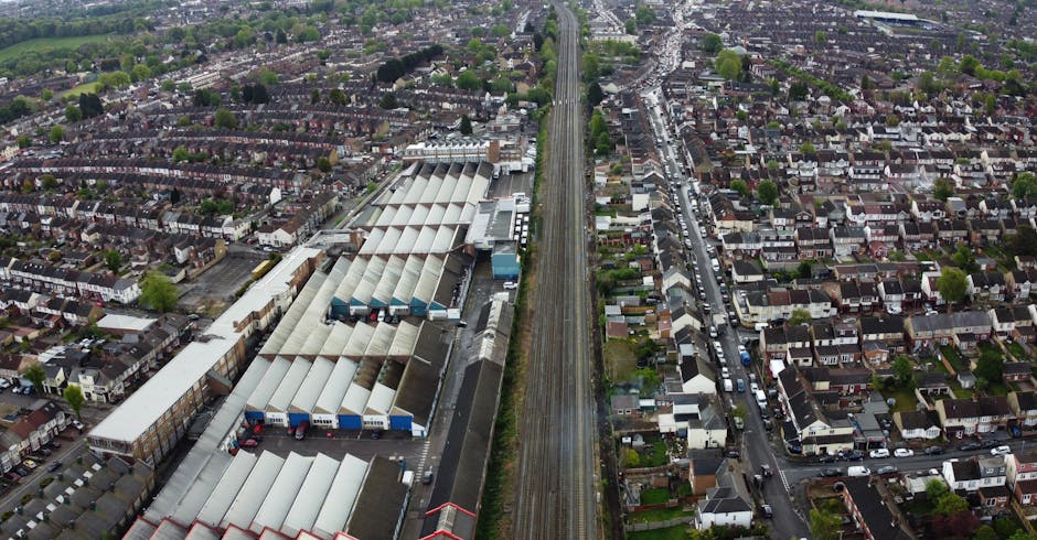 Aerial view of a residential area adjacent to a large rail freight depot situated along a straight railway line, with multiple industrial warehouses featuring metal roofs on the left side, and rows of terraced houses with small gardens and narrow streets on the right. The scene is captured during daylight with clear visibility of parked cars on the streets and the surrounding greenery, illustrating an urban environment suitable for house removals and moving services by companies like Man with Van Upminster. The image emphasizes the proximity of residential properties to transport infrastructure, relevant for planning home relocation and furniture transport logistics.
