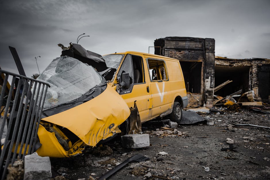 A yellow panel van with significant front-end damage, including a crushed and deformed hood and windshield, sits amidst debris and rubble in front of a partially destroyed brick building. The van appears to have been involved in a severe collision or explosion, with the surrounding ground covered in scattered concrete blocks, broken wood, and other building materials. There are no visible occupants or movement in the scene. Nearby, a metal fence is bent and partially toppled, and the sky above is overcast with dark clouds, creating a somber atmosphere. This scene depicts the aftermath of a destructive event, with the van positioned at the center of the damage, highlighting the extent of the destruction. The background building shows extensive fire and structural damage, with blackened walls and collapsed sections, illustrating the severity of the incident, relevant to the context of removals, damage assessment, and logistical challenges faced after such destructive events. Man with Van Upminster specializes in handling complex recovery and house removal scenarios, including damage claims and transport of salvageable items amidst difficult circumstances.
