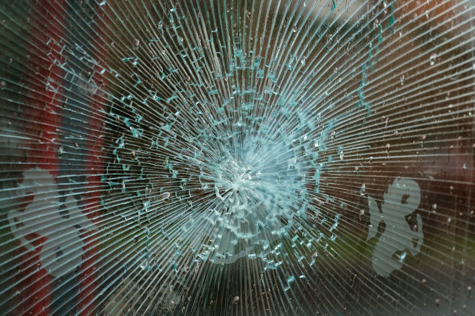 Close-up view of a shattered glass window with a large, central impact point radiating cracks outward in multiple directions. The glass fragments are irregular, with some shards remaining attached around the edges of the impact. The background behind the glass is blurred, showing hints of red and green hues, indicative of outdoor or indoor surroundings. The image captures the detail and texture of the broken glass surface, highlighting the results of accidental damage that can occur during house removals or furniture transport. This detailed visual underscores the importance of careful handling and packing during home relocation, as managed by companies like Man with Van Upminster, especially when dealing with fragile objects such as glass.