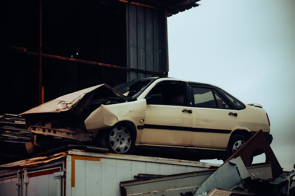 A yellow panel van with significant front-end damage, including a crushed and deformed hood and windshield, sits amidst debris and rubble in front of a partially destroyed brick building. The van appears to have been involved in a severe collision or explosion, with the surrounding ground covered in scattered concrete blocks, broken wood, and other building materials. There are no visible occupants or movement in the scene. Nearby, a metal fence is bent and partially toppled, and the sky above is overcast with dark clouds, creating a somber atmosphere. This scene depicts the aftermath of a destructive event, with the van positioned at the center of the damage, highlighting the extent of the destruction. The background building shows extensive fire and structural damage, with blackened walls and collapsed sections, illustrating the severity of the incident, relevant to the context of removals, damage assessment, and logistical challenges faced after such destructive events. Man with Van Upminster specializes in handling complex recovery and house removal scenarios, including damage claims and transport of salvageable items amidst difficult circumstances.