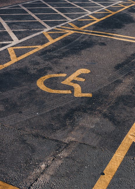 Close-up view of an asphalt parking lot featuring yellow painted lines and a national wheelchair symbol indicating accessible parking. The surface appears worn with visible patches and textured markings, and the concrete curb edge is partially visible at the bottom right. The parking space is situated outdoors with natural lighting. This setting is typical for residential or commercial relocations, where access routes and parking arrangements play a crucial role in the logistics of home removal or furniture transport. For professional removals in Upminster, Man with Van Upminster offers reliable services that coordinate loading and unloading processes in accessible locations like this. The image emphasizes the importance of designated parking areas for efficient and safe moving operations during packing and moving activities.