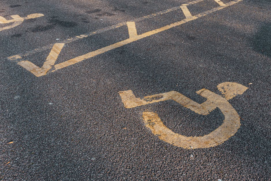 Close-up view of an asphalt parking lot featuring yellow painted lines and a national wheelchair symbol indicating accessible parking. The surface appears worn with visible patches and textured markings, and the concrete curb edge is partially visible at the bottom right. The parking space is situated outdoors with natural lighting. This setting is typical for residential or commercial relocations, where access routes and parking arrangements play a crucial role in the logistics of home removal or furniture transport. For professional removals in Upminster, Man with Van Upminster offers reliable services that coordinate loading and unloading processes in accessible locations like this. The image emphasizes the importance of designated parking areas for efficient and safe moving operations during packing and moving activities.