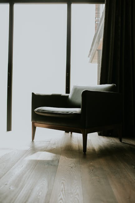 A black armchair with light-colored cushions is positioned inside a room near large floor-to-ceiling windows, which are covered partially by dark curtains. The armchair is placed on a wooden floor with natural light illuminating the space, indicating daytime. In the background, part of a balcony or exterior building structure is visible through the windows. The room appears to be in the process of a home relocation or furniture transport, with the setting prepared for packing or moving services by Man with Van Upminster, a company specialising in removals and furniture transport, evident from the professional arrangement of the space and the focus on interior details relevant to moving logistics.
