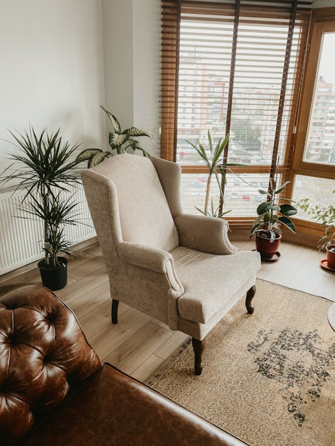 A beige upholstered armchair with dark wooden legs positioned near a large window with wooden blinds in a living room. The room features light-colored wooden flooring, a light-colored wall with a radiator, and several potted houseplants, including a tall leafy plant in a black pot and a smaller plant in a reddish-brown pot. In the foreground, part of a brown leather Chesterfield sofa is visible, and a beige patterned rug partially covers the floor. The scene appears well-lit by natural daylight, illustrating a typical setting where furniture may be prepared for a home relocation or moving process, as handled by Man with Van Upminster, specialists in removals and furniture transport.