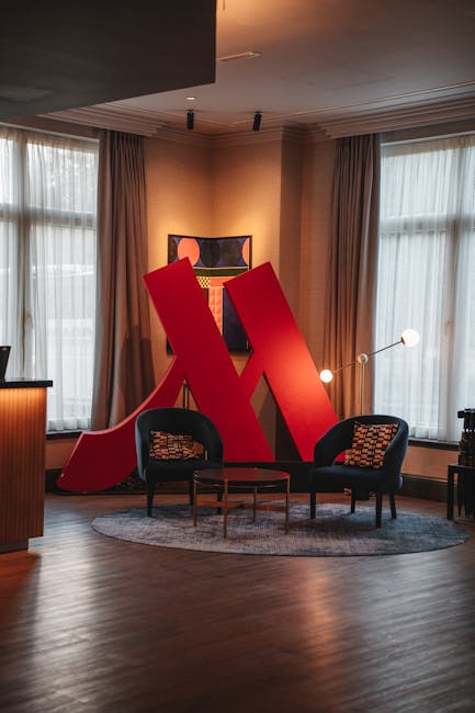 A black armchair with light-colored cushions is positioned inside a room near large floor-to-ceiling windows, which are covered partially by dark curtains. The armchair is placed on a wooden floor with natural light illuminating the space, indicating daytime. In the background, part of a balcony or exterior building structure is visible through the windows. The room appears to be in the process of a home relocation or furniture transport, with the setting prepared for packing or moving services by Man with Van Upminster, a company specialising in removals and furniture transport, evident from the professional arrangement of the space and the focus on interior details relevant to moving logistics.
