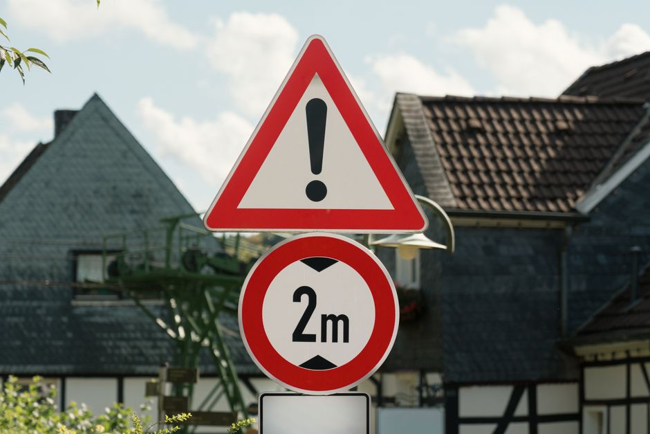 A photograph of two traffic signs mounted on a metal pole in front of residential houses with dark tiled roofs and brick or weatherboard facades. The upper sign is a red-bordered triangle with a black exclamation mark inside, indicating a general warning. Below it is a circular sign with a red border and a white background displaying '2m,' indicating a height restriction of two meters. The signs are positioned on a sidewalk or pavement area, with part of a green tree branch visible on the left. In the background, houses with pitched roofs and small windows are partially obscured by the signs and sky, which features some scattered clouds. The scene appears to be in a quiet residential neighbourhood, relevant to the context of house removals, storage, or parking restrictions during a home relocation, as illustrated by the image and supported by the services offered by Man with Van Upminster.