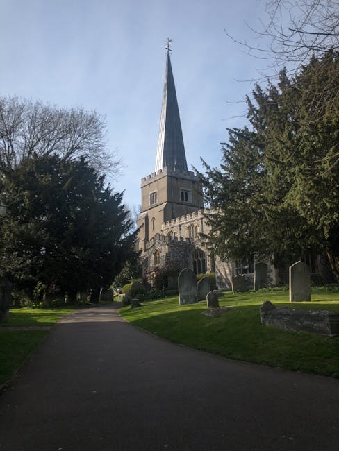 A paved pathway leading through a churchyard with several gravestones and mature trees on either side, in front of a historic stone church with a tall, pointed spire extending into a partly cloudy sky. The church is framed by greenery, and sunlight casts shadows on the grass and pathway. This scene illustrates the exterior environment of a traditional UK place of worship, highlighting the architectural features, the surrounding landscape, and the natural light conditions, which are relevant for home relocation and loading processes managed by companies like Man with Van Upminster during moving services.