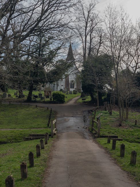 A paved pathway leading through a grassy area with wooden post barriers lining each side, situated within a park or green space next to a historic windmill, with leafless trees in the foreground and a church with a tall steeple visible in the background on a cloudy day. This setting depicts an outdoor environment typical for residential or community grounds in Upminster, suitable for moving preparations or outdoor storage during a home relocation. The scene may be part of a moving route or landscape involved in furniture transport, aligning with house removals services offered by Man with Van Upminster. The image captures the natural surroundings and pathway used for transporting boxes, furniture, or equipment during a household move in tight street or outdoor environments, illustrating the logistical aspects of a home relocation in Upminster.