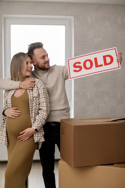 Inside a home during a house removal, a smiling couple stands next to a large cardboard box on the floor, which is partially unsealed and ready for packing or transport. The man is holding a rectangular sign with red 'SOLD' lettering, indicating a completed property sale. The woman, visibly pregnant, is wearing a beige dress and checked shirt, while the man is dressed in a light-colored sweater and dark trousers. They are positioned near a window that allows natural light into the room, highlighting grey painted walls and a clean, minimal interior. Behind them, a plain, light-colored wall and a window with simple framing are visible. The scene captures a moment of moving or packing, relevant to professional removals and home relocation services, such as those offered by Man with Van Upminster, emphasizing the process of preparing a property for sale or ownership transfer.