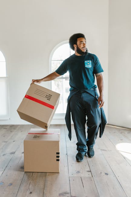 A man with dark curly hair and a beard, dressed in a navy blue t-shirt and dark trousers, is inside a bright, empty room with white walls and a large arched window in the background. He is holding a cardboard box wrapped with red tape in his right hand, appearing to be in the process of loading or unloading it. On the light wooden floor, there is another cardboard box with printed labels and black tape, positioned directly below the first box. The room has natural light coming through the window, illuminating the scene, which suggests a home relocation or furniture transport process. This image showcases a professional moving service, such as Man with Van Upminster, involved in packing and preparing items for moving, reflecting aspects of home removals involving careful handling of boxes during a loading or unloading stage.