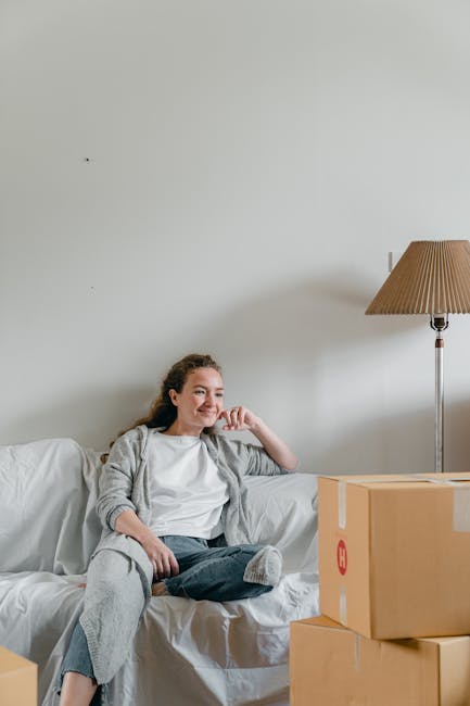 Inside a home during a house removal, a smiling couple stands next to a large cardboard box on the floor, which is partially unsealed and ready for packing or transport. The man is holding a rectangular sign with red 'SOLD' lettering, indicating a completed property sale. The woman, visibly pregnant, is wearing a beige dress and checked shirt, while the man is dressed in a light-colored sweater and dark trousers. They are positioned near a window that allows natural light into the room, highlighting grey painted walls and a clean, minimal interior. Behind them, a plain, light-colored wall and a window with simple framing are visible. The scene captures a moment of moving or packing, relevant to professional removals and home relocation services, such as those offered by Man with Van Upminster, emphasizing the process of preparing a property for sale or ownership transfer.
