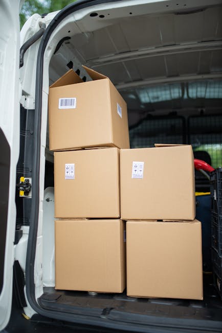 Inside the rear compartment of a white van used by Man with Van Upminster for home relocation, four large cardboard boxes are stacked, with two positioned on the floor and two placed on top. The boxes are secured with plastic wrap and have labels indicating they contain household items. The van's interior is illuminated by natural light, revealing a dark interior with visible metal framework and a black trolley or cart to the right side, which is used for furniture transport and packing during the loading process. The background includes part of the vehicle's black interior paneling and a glimpse of the outside environment through the van's open door. This scene captures the logistics involved in furniture transport and moving preparations, consistent with professional removals services referenced in the webpage about van loading bays and parking suspensions in RM14, Upminster.