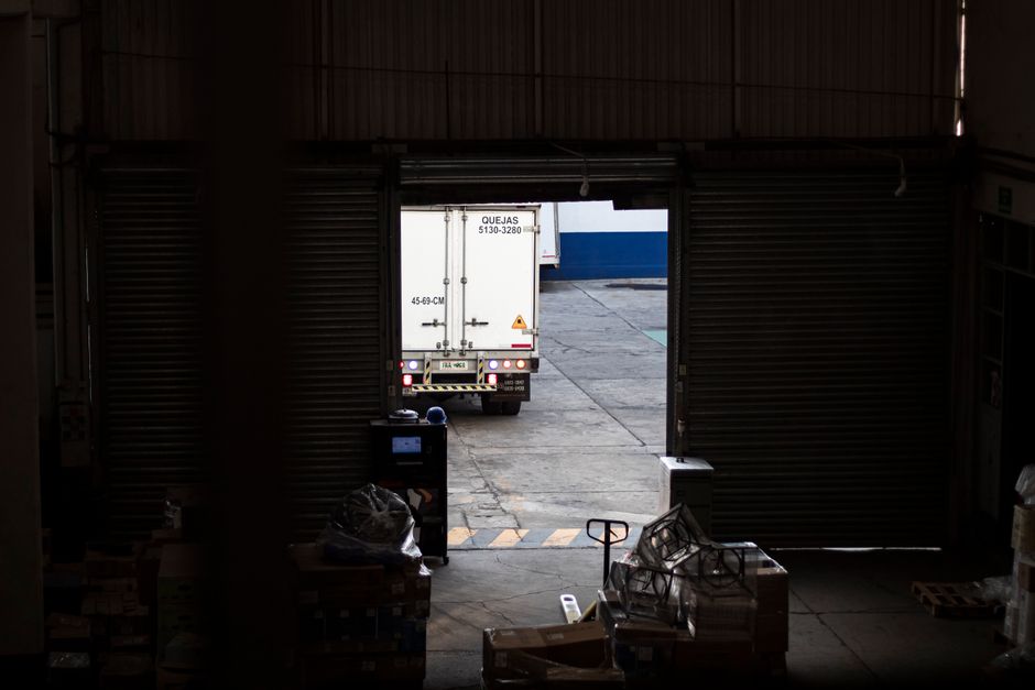 View from inside a warehouse or storage facility looking out through an open roller shutter door towards an outdoor loading area with a white moving truck parked on a paved surface. The truck's rear doors are closed, displaying contact details and size specifications. Inside the warehouse, various cardboard boxes, plastic-wrapped items, and packing materials are stacked and scattered near the entrance, indicating ongoing packing or furniture transport activities. A trolley and other moving equipment are positioned near the doorway, emphasizing preparation for customer house removals or home relocation tasks. The scene depicts a professional moving process being carried out by [COMPANY_NAME], focusing on loading furniture and boxes into the truck, with a subtle connection to removals, packing, and transport logistics in the context of house moving services. Natural daylight illuminates the scene outside, contrasting with the darker interior.