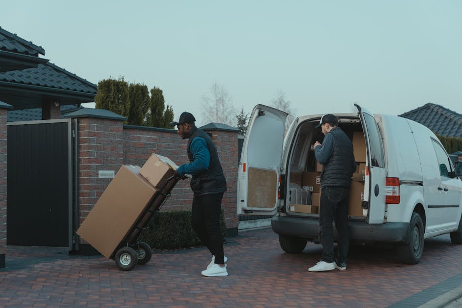 Inside the rear compartment of a white van used by Man with Van Upminster for home relocation, four large cardboard boxes are stacked, with two positioned on the floor and two placed on top. The boxes are secured with plastic wrap and have labels indicating they contain household items. The van's interior is illuminated by natural light, revealing a dark interior with visible metal framework and a black trolley or cart to the right side, which is used for furniture transport and packing during the loading process. The background includes part of the vehicle's black interior paneling and a glimpse of the outside environment through the van's open door. This scene captures the logistics involved in furniture transport and moving preparations, consistent with professional removals services referenced in the webpage about van loading bays and parking suspensions in RM14, Upminster.